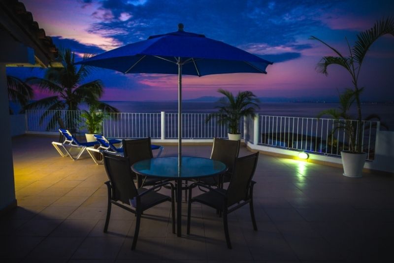 A rooftop patio dining setting with a blue umbrella overlooking the ocean at dusk