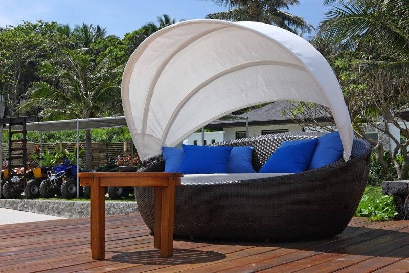 A wicker outdoor daybed with a canopy shade and blue cushions on a timber deck
