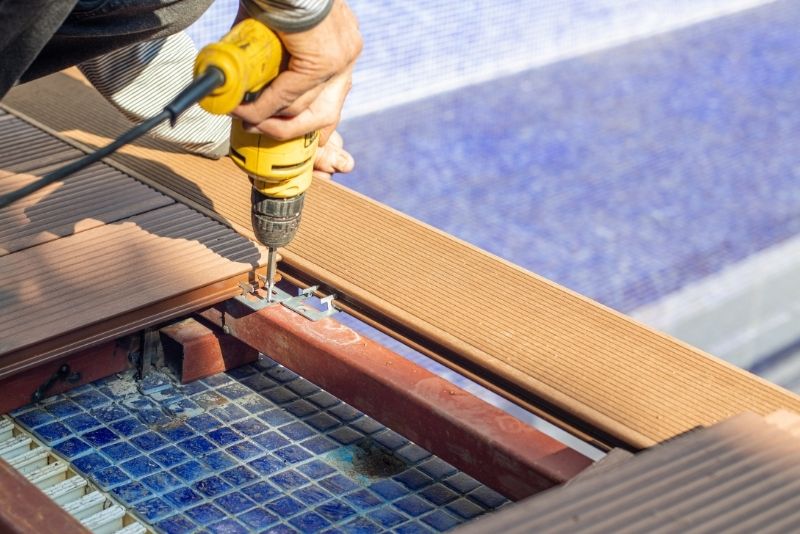 Tradesperson installing composite decking boards with a drill beside a pool