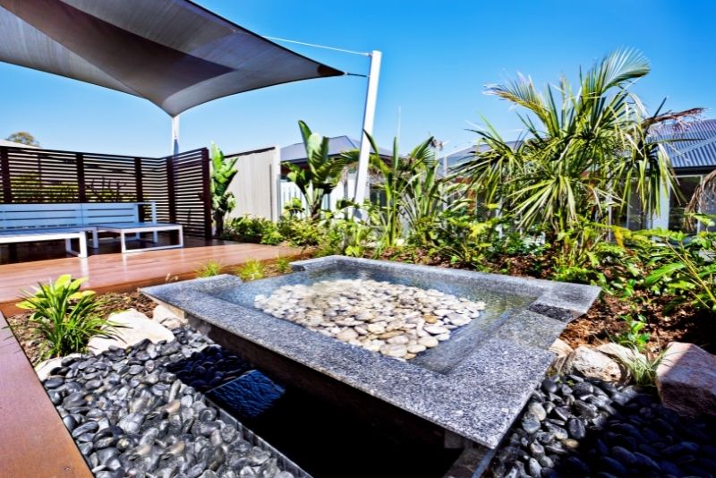 A shade sail over a timber deck with a stone water feature and tropical landscaping